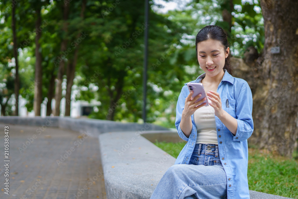 Fototapeta premium Smiling female student using smartphone sitting in campus space near university park. asian college student chats online, browses social networks reads messages, scroll looking at phone screen