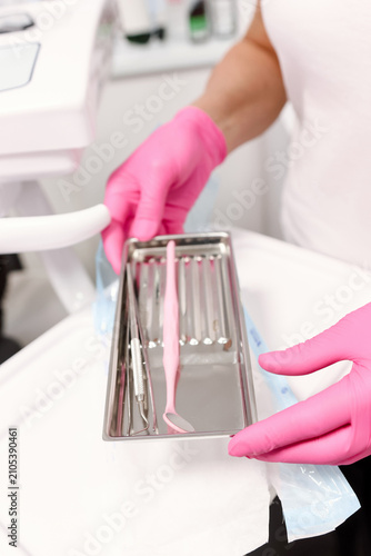Dentist wearing pink gloves holds a sterile metal tray with a dental mirror and examination instruments before treatment.