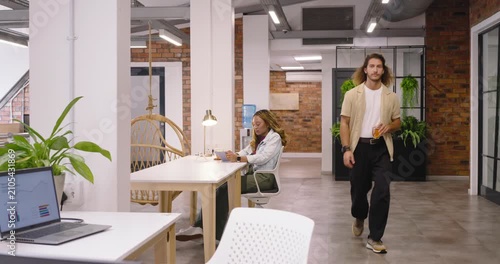 Diverse team in open office man entering with orange juice, pulling chair, powering laptop, typing