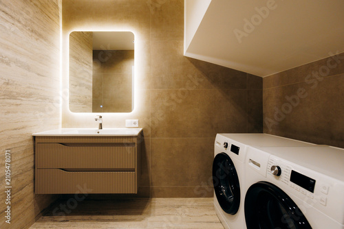 Modern laundry bathroom with illuminated mirror, floating vanity, front load washer and dryer beneath sloped ceiling, warm neutral finishes and compact storage, with bathroom, laundry, washer, dryer