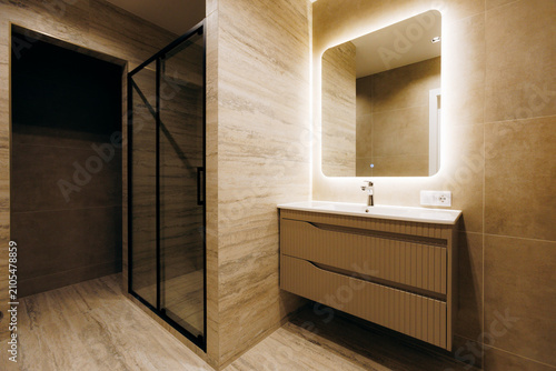 Modern hotel bathroom with illuminated mirror, floating vanity, glass shower enclosure, warm neutral tones, wall mounted faucet, ribbed storage, clean contemporary interior