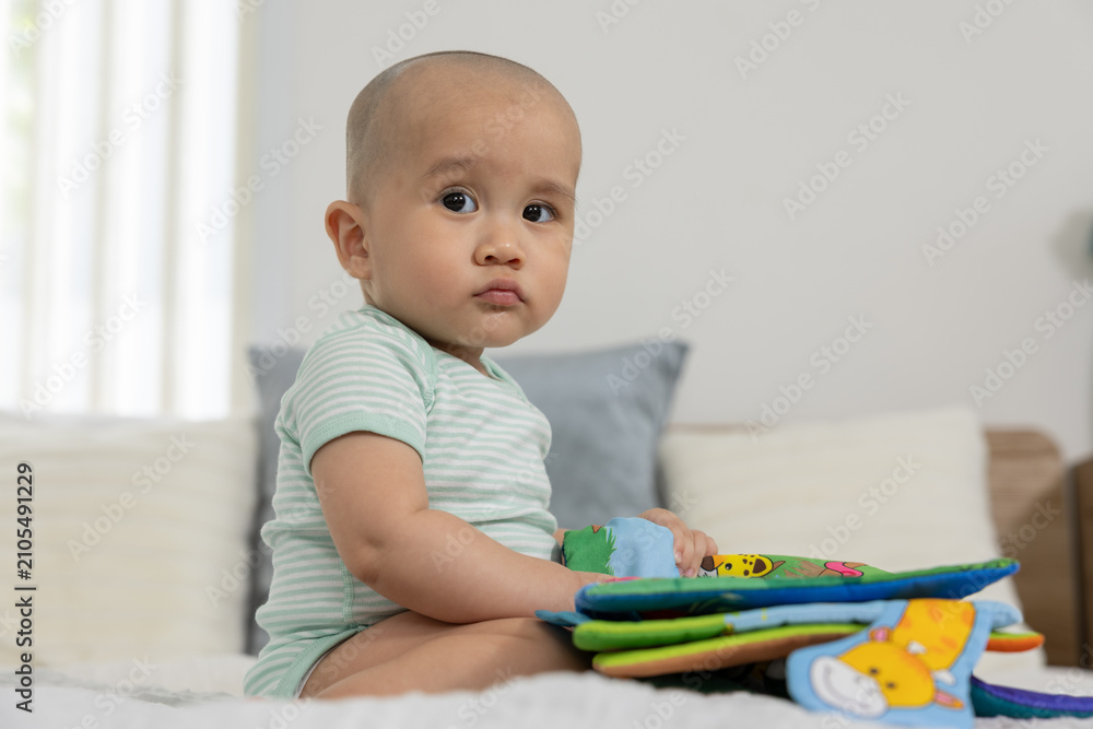 Naklejka premium Baby infant toy book bed bedroom child cute curious baby sitting on bed holding colorful soft fabric book in bright bedroom with pillow and natural light calm curious expression