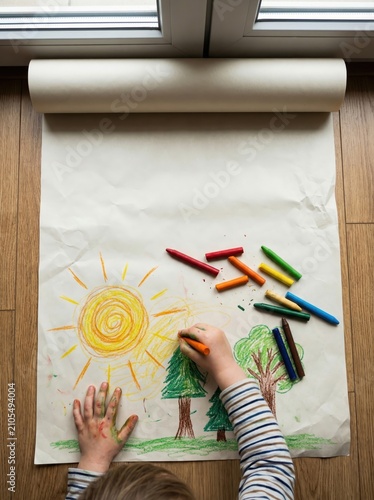 top view of caucasian child hands drawing bright sun and trees with colorful crayons on large paper roll on wooden floor. childhood creativity concept. poster. copyspace.