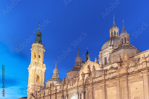 Illuminated Cathedral-Basilica of Our Lady of the Pillar in Zaragoza at night. Beautiful baroque architecture with towers and domes under a deep blue twilight sky in Spain