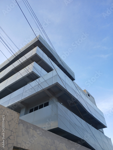 Low angle view of a sleek modern commercial building facade with metallic paneling and geometric architectural lines set against a clear blue sky during a bright and sunny afternoon day.