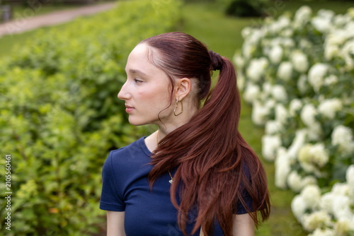 Fotografie Red-haired teenage girl touches her long hair while walking through blooming park on warm summer evening