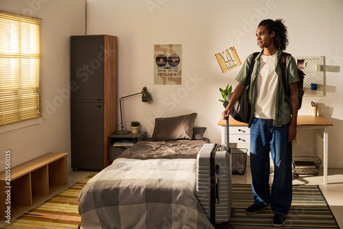 Young adult man standing with suitcase in dorm room, arriving for college move in and starting independent student life. Useful for education marketing, housing, orientation campaigns