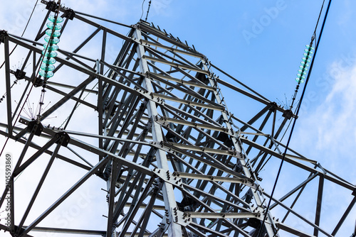 High-voltage metal power transmission pylon against a clear blue sky, showing complex steel framework, ceramic glass insulators and conductive cables in a low angle architectural perspective