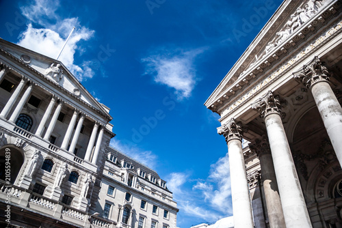 City of London financial district. Historic neoclassical pillars frame the Bank of England exterior under a clear blue sky.