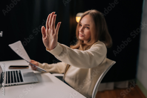 Portrait of serious confident businesswoman focused at desk holding papers and extends hand in stop gesture to signal refusal of interruption working on laptop in dark modern office room.