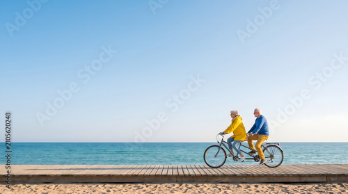 Elderly couple riding a tandem bicycle on a wooden boardwalk by the seaside