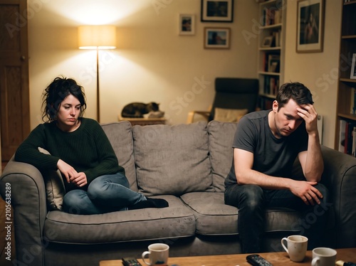 Couple sitting on the couch feels disconnected while experiencing emotional distance in their dark room.