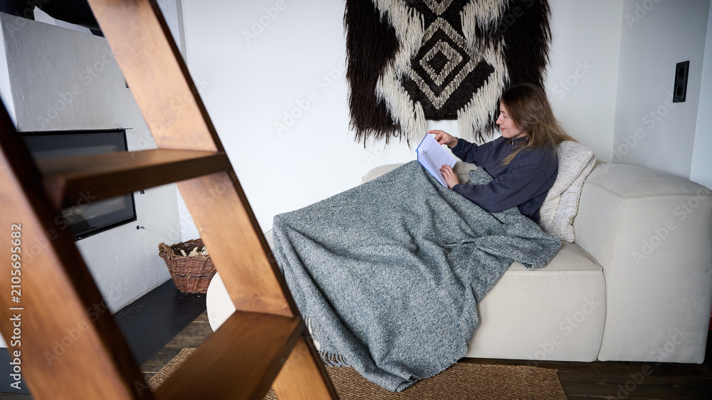 Obraz premium Woman reading book on sofa wrapped in blanket, relaxing at home near fireplace, cozy interior with warm atmosphere, leisure time and comfortable lifestyle concept