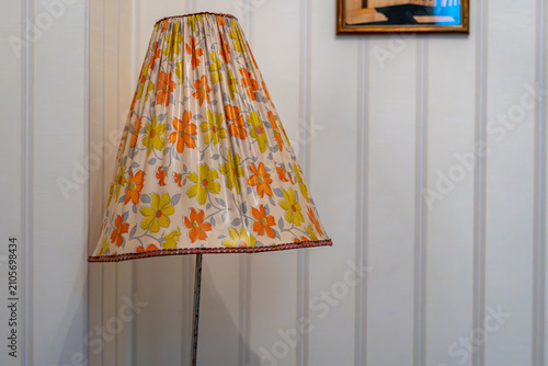 Photography Vintage floor lamp from the PRL era with a conical lampshade featuring a retro floral pattern in shades of orange and yellow, positioned against a light wooden paneled wall, interior design