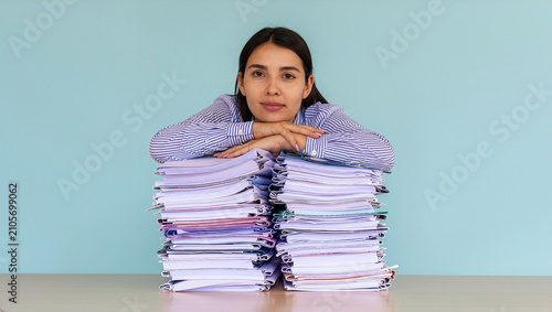 A tired businesswoman leaning her arms and chin on two high stacks of paper folders on a desk against a plain blue background, depicting office exhaustion, heavy workload, and corporate stress.