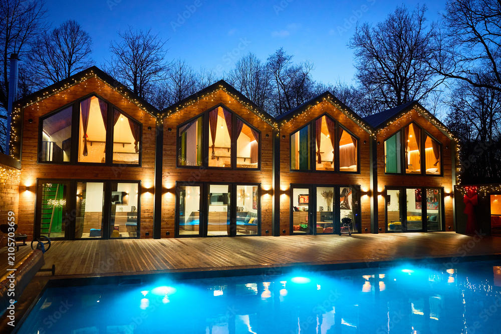 Obraz premium Modern spa resort with glowing wooden lodges and bright pool at night. Cozy design and serene setting illustrate hospitality service, leisure lifestyle and high end holiday concept