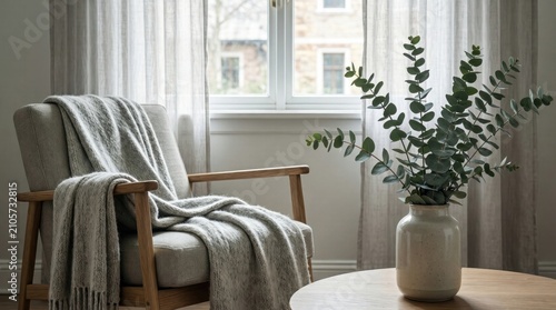 A chair with a blanket draped over it sits by a window. A plant in a vase is on a table nearby. The room is bright and has soft curtains.