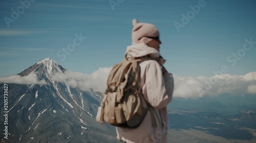 Young beautiful girl wearing sunglasses and carrying backpack stands against backdrop of majestic volcano and admiring surroundings from above.