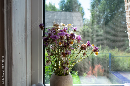 Quadro su tela Dried wildflowers arranged in a ceramic vase sit on a sunlit windowsill, casting a soft still-life mood against a rustic garden backdrop