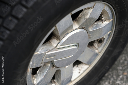 close‑up shows a damaged silver alloy wheel with fractured spokes, chipped edges, and a central stylized emblem; the adjacent tire tread and rim edge highlight the impact damage