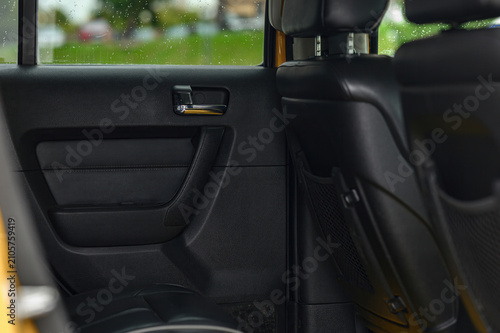 car rear door panel: matte black textured trim, a metallic door handle with chrome accents, a mesh speaker grille, and part of the black leather rear seat with yellow trim