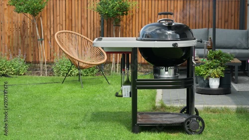 A closed charcoal grill steams on a green lawn in a modern backyard patio terrace. Light smoke drifts from the kettle grill during a summer barbecue cookout. Great setting for outdoor dining.