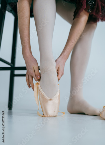 The photo shows a ballerina's legs wearing pointe shoes during ballet practice or performance. Close-up view of classical dance footwear, indoor studio or stage setting, professional dance attire.
