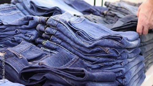 Customer choosing blue denim jeans from folded stacks on display in clothing store, close-up shopping for casual fashion apparel in retail