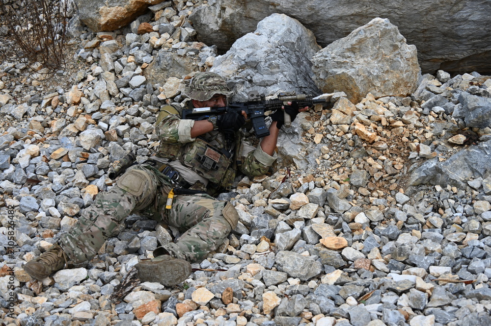Obraz premium Focused military infantry soldier lying prone on rugged rocky terrain aiming a tactical assault rifle during a dangerous combat training mission for national defense and special security.