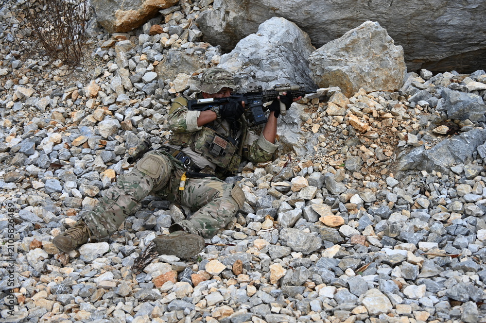 Obraz premium Focused military infantry soldier lying prone on rugged rocky terrain aiming a tactical assault rifle during a dangerous combat training mission for national defense and special security.