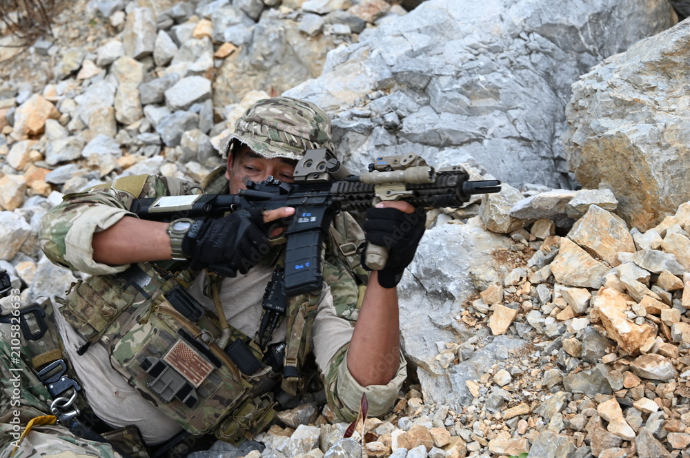 Obraz premium Focused military infantry soldier lying prone on rugged rocky terrain aiming a tactical assault rifle during a dangerous combat training mission for national defense and special security.