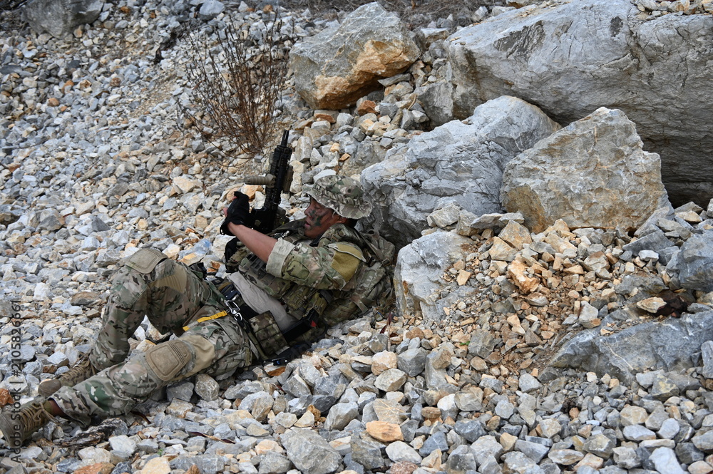 Obraz premium Focused military infantry soldier lying prone on rugged rocky terrain aiming a tactical assault rifle during a dangerous combat training mission for national defense and special security.