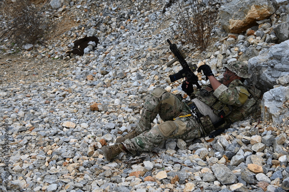 Obraz premium Focused military infantry soldier lying prone on rugged rocky terrain aiming a tactical assault rifle during a dangerous combat training mission for national defense and special security.