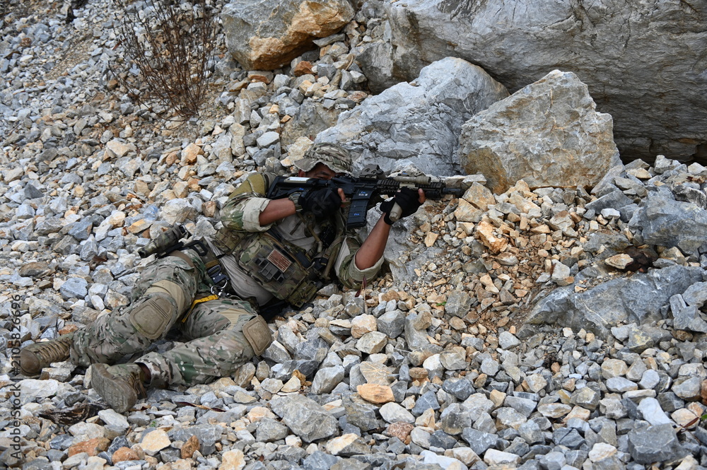 Obraz premium Focused military infantry soldier lying prone on rugged rocky terrain aiming a tactical assault rifle during a dangerous combat training mission for national defense and special security.
