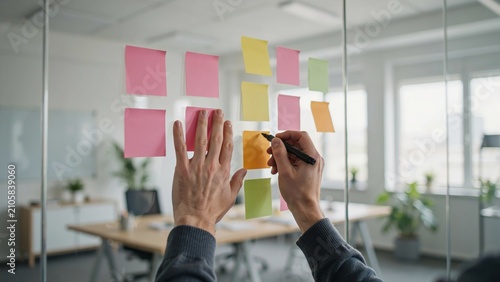 Close-up of hands writing on colorful sticky notes stuck to glass office wall, pink yellow green and orange memo papers with black marker pens, blurred desks and plants in modern workspace background