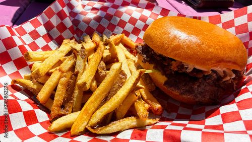 A hamburger topped with brisket and served with fries, in a basket with a red a white checked liner. Seved outside on a picnic table.