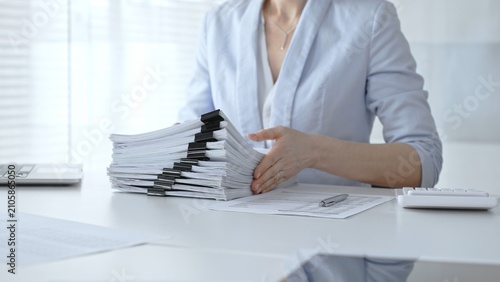 Obraz na plátně Businesswoman's hands organizing a towering stack of bound reports and financial documents on a white desk, navigating paperwork overload in a busy corporate office environment