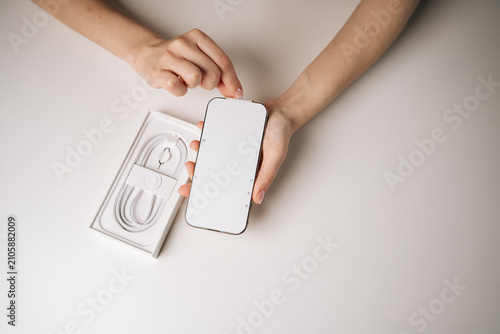 Top view of woman unpacking new smartphone and removing screen protector sitting on white table, with charging cable in box visible in background. Concept of excitement of unboxing new device.