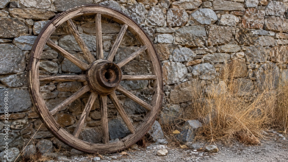 Obraz premium Weathered wooden wagon wheel leaning against a dry stone wall with gravel and dried grasses