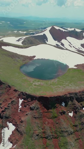 Vertical Drone Shot of Volcanic Crater Lake surrounded by Red Soil, Green Slopes, and Melting Snow