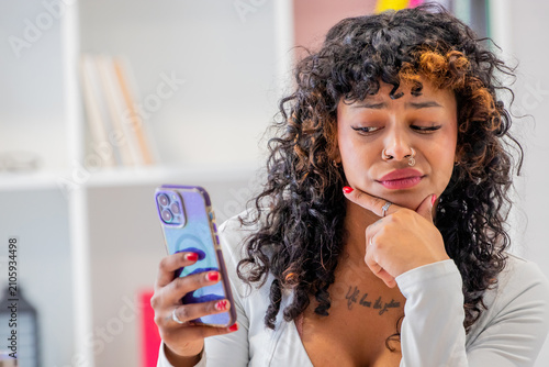  young latina woman with curly hair sits at home, looking skeptically at her smartphone, holding a confused, doubtful expression while reading a message