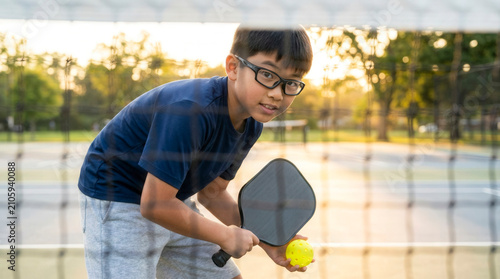 Asian kid playing ball with pickleball paddle on outdoor court, youth sports training and healthy active lifestyle