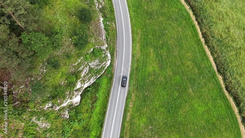 Aerial view of a black car driving along a winding rural road surrounded by lush green trees and rocky cliffs. Travel by auto at scenic landscape. Vehicle journey through natural beauty of countryside