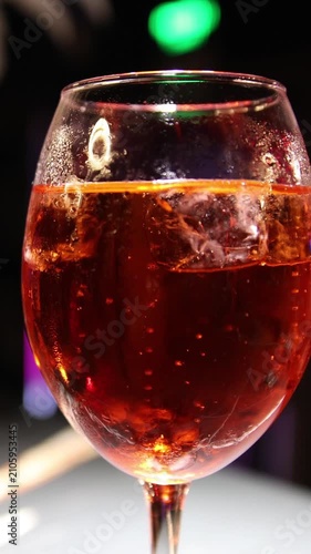 Vibrant Aperol Spritz cocktail with ice cubes and condensation droplets in stemmed glass against dark bar background with blurred glowing green bokeh light.