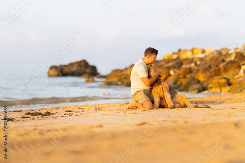 Bild auf Leinwand Young man kneeling on sandy beach embraces golden retriever dog with rocky shoreline and calm ocean waves visible in the background during sunset