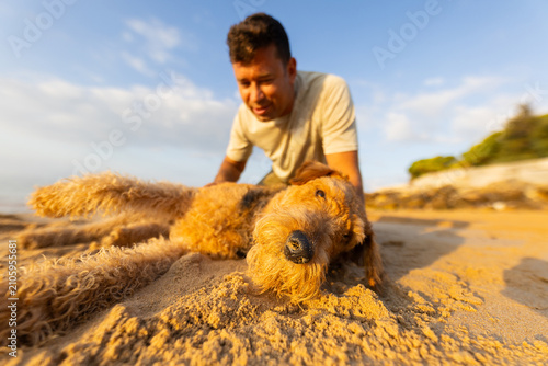Photos Man playing with a brown dog on sandy beach during sunset, with ocean waves and green vegetation visible in the background