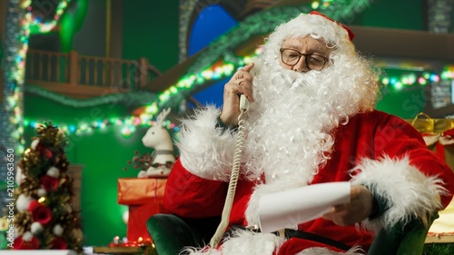 Photography Santa Claus answers a landline phone call and talks with children requesting Christmas gifts, responding to their wishes while dressed in a festive costume during the holiday season