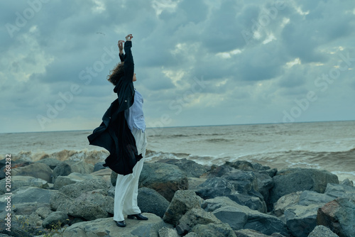 фотографія Woman stands on rocky shore with arms raised in celebration, wind whipping cloth