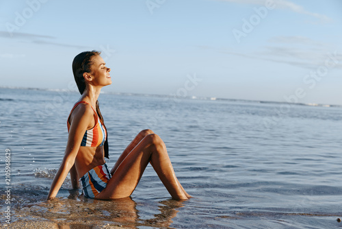Slika na platnu Young woman in a striped bikini sits at the edge of the calm sea, eyes closed, basking in warm sun and a gentle breeze, relaxed and content