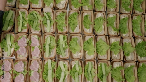 Food preparation process featuring a person assembling sandwiches with lettuce, ham, and bread in a commercial kitchen setting, showcasing organized trays of ingredients